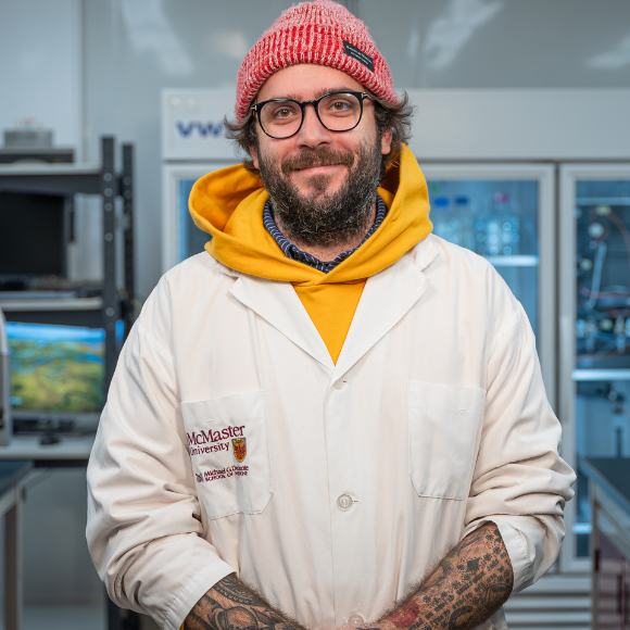 John Stokes, a bearded tattooed man in a hoodie and a lab coat, with glasses and a toque, smiling in his lab.