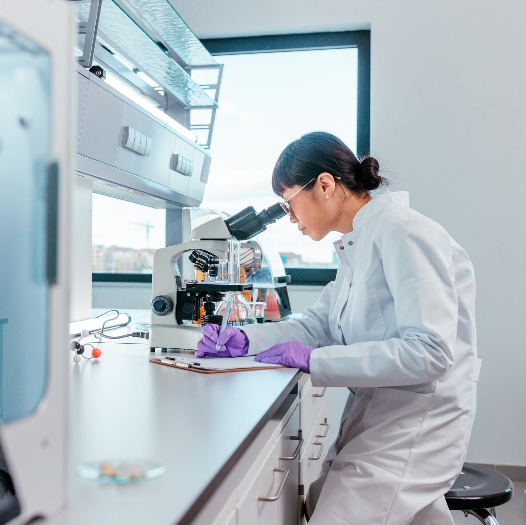 A female scientist is sitting in her lab and looking trough the microscope, she is writing down notes about her discoveries after looking at samples.