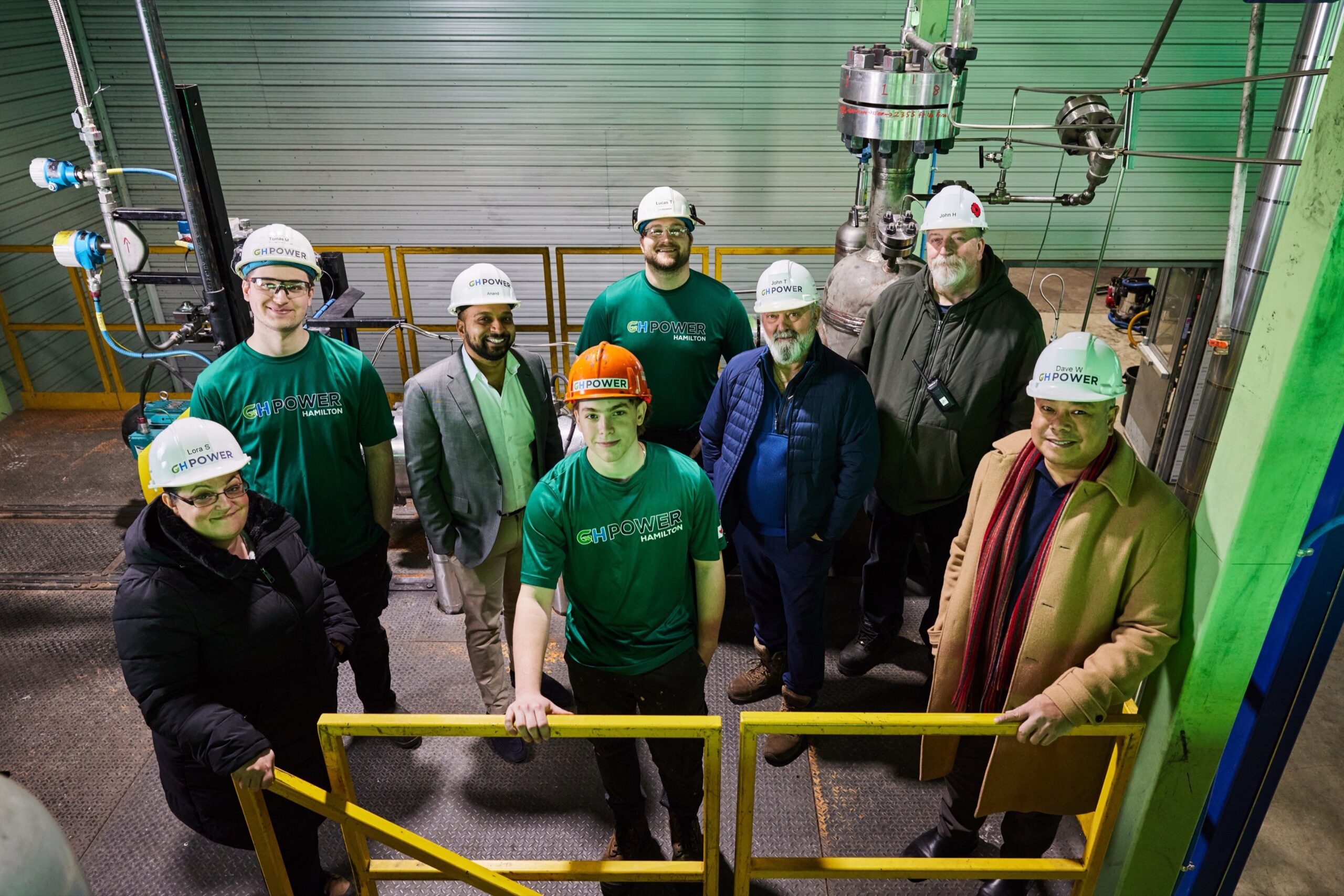 Smiling group of 8 staff members in plant wearing hardhats
