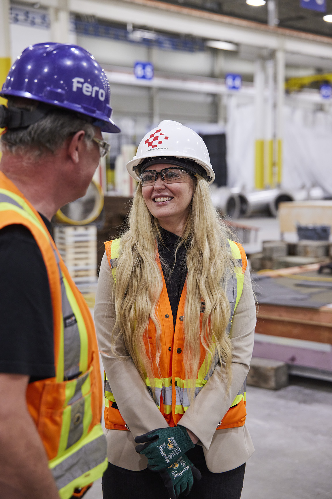 Sabrina Fiorellino - Blonde woman smiling in hardhat speaking with worker in orange vest, in manufacturing plant