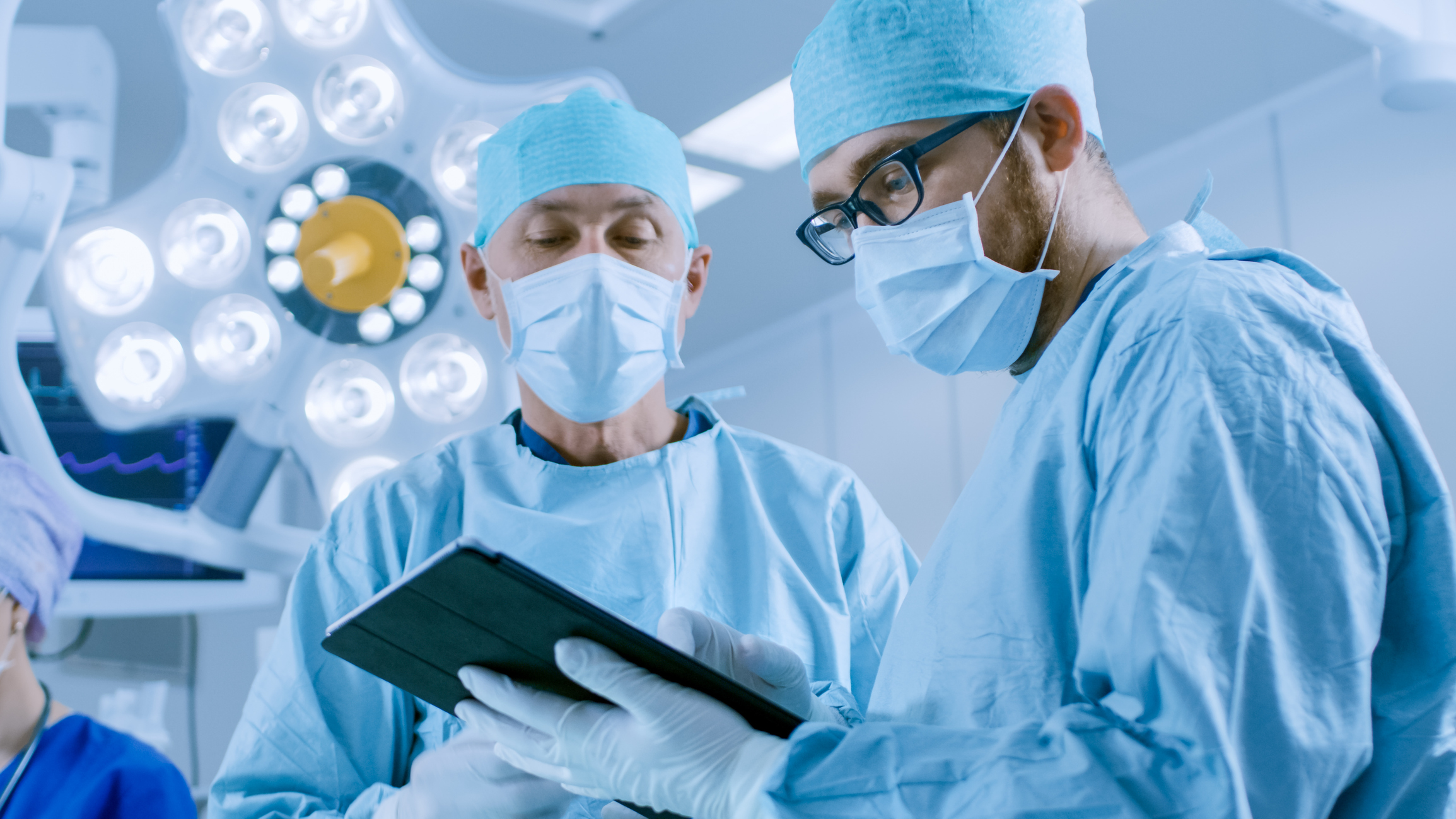 Two Professional Surgeons Use Digital Tablet Computer while Standing in the Modern Hospital Operating Room